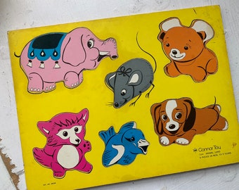 May include: A colorful wooden puzzle featuring six animal shapes: a pink elephant, a gray mouse, an orange bear, a brown and white dog, a pink fox, and a blue bird. The puzzle pieces are on a yellow background with the text "Connor Toy 11/4 Animal Land 6 Pieces 18 Mos. To 5 Years" printed on the bottom right corner.
