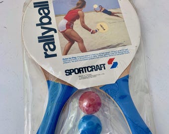 May include: A set of two blue and wooden rallyball paddles with two balls, one red and one blue. The packaging includes instructions for play and the text "Carry bag, two paddles, two balls for lawn or beach play, fun for all ages." and "rallyball".