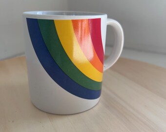 May include: White ceramic mug featuring a vibrant rainbow design. The rainbow arcs across the mug in bands of red, orange, yellow, green, and blue. The mug has a simple, curved handle and is set on a light-colored wooden surface.