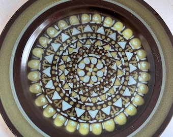 May include: A round ceramic plate featuring a dark brown rim and a central circular pattern. The design consists of concentric rings of light blue and pale yellow geometric shapes, including triangles, circles, and stylized floral motifs.