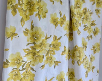 May include: White fabric with a repeating pattern of large yellow flowers and green leaves. The fabric is hanging from a wooden rod, showcasing the floral design. The flowers have multiple layers of petals.