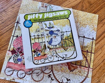 May include: A Jiffy Jigsaws 144-piece interlocking puzzle box measuring 12 inches by 12 inches, featuring an illustration of a person on a bicycle with a wagon, placed atop the completed puzzle showing the same scene with a black fence.