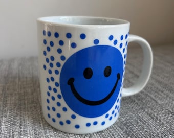 May include: White ceramic mug with a blue smiley face and blue polka dots. The mug has a curved handle and a simple, clean design. The smiley face is centered on the mug, with the polka dots scattered around it.