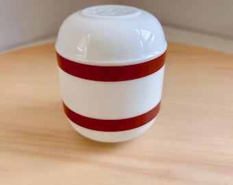 May include: A white, egg-shaped salt shaker with two horizontal red bands. The top of the shaker has small holes for dispensing salt. The shaker is sitting on a light-colored wooden surface.