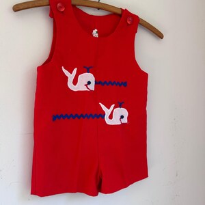 May include: Red romper with white whale appliqués and blue wave accents. The sleeveless romper has red buttons on the shoulders and is hanging on a wooden hanger. A vintage children's garment.