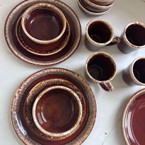 May include: A collection of brown ceramic dinnerware, including plates, bowls, and mugs. The plates and bowls have a dark brown center with a lighter brown and cream-colored rim. The mugs are a similar brown color.