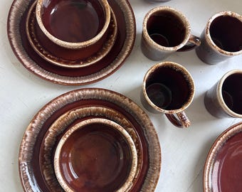 May include: A collection of brown ceramic dinnerware, including plates, bowls, and mugs. The plates and bowls have a dark brown center with a lighter brown and cream-colored rim. The mugs are a similar brown color.