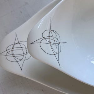 May include: Two white ceramic serving dishes with a unique, abstract black line design. The dishes have a smooth, glossy finish and a slightly curved, elongated shape. The black design is a series of intersecting lines and shapes.