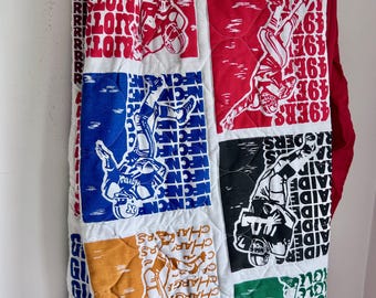 May include: A quilted blanket featuring various football team logos and player illustrations in red, blue, black, green, and gold. The quilt has a white background with text such as "49ers", "Colts", and "Chargers".
