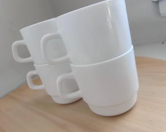 May include: A stack of four white ceramic mugs with rounded handles. The mugs are stacked on top of each other, with the top mug slightly angled. The mugs are on a light brown wooden surface.