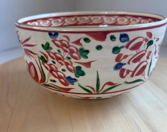 May include: A cream-colored ceramic bowl with a red, green, and blue floral design. The rim has a red border with a gold accent. The bowl is sitting on a light-colored wooden surface.