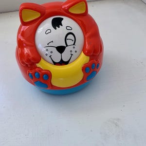 May include: A red, yellow, and blue plastic toy shaped like a cat. The cat has a white face with black eyes and a black nose. The toy has a yellow ring around the middle and blue paw prints on the bottom.