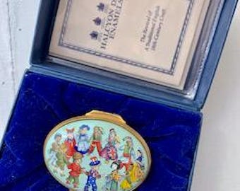 May include: A blue and white oval enamel box with a gold trim. The box features a colorful scene of children playing. The box is in a blue velvet box with a white card that reads "Halcyon Days Enamels".