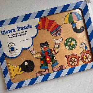 May include: A wooden Clown Puzzle by Discovery Toys featuring a central clown figure surrounded by a bicycle, umbrella, accordion, and horn. The puzzle pieces have yellow knobs and are housed in a blue and white striped cardboard frame.
