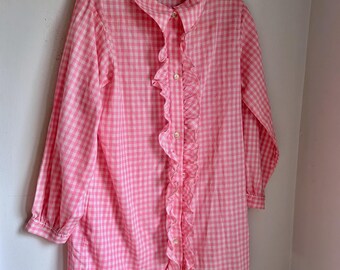May include: Pink and white gingham patterned shirt with a ruffled front and button closure. The long-sleeved shirt is hanging on a red hanger. The shirt has a classic collar and a relaxed fit.