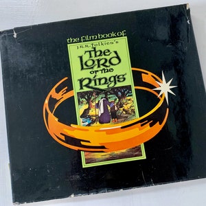 May include: A black box with a gold ring and a green and white image of a scene from "The Lord of the Rings" movie. The box has the text "the film book of J.R.R. Tolkien's The Lord of the Rings" on the front.