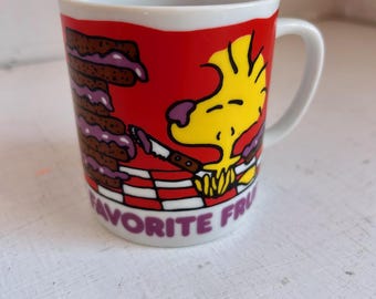 May include: A white ceramic mug with a red background featuring a yellow bird character holding a knife, a stack of brown toast with purple jam, and a red and white checkered pattern. The text FAVORITE FRU is printed in purple at the bottom.