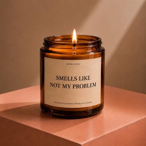 May include: A lit candle in an amber glass jar with a beige label. The label reads AMBER CANDLE, SMELLS LIKE NOT MY PROBLEM, and 100% SOY WAX HAND POURED IN CANADA. The candle features a single burning wick centred within the wax inside the jar.