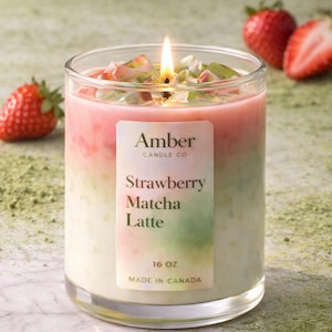 May include: A lit candle in a clear glass jar with a pink and green layered design. The label reads "Amber Candle Co. Strawberry Matcha Latte 16 OZ Made in Canada." Fresh strawberries are in the background.