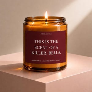 Peut inclure: Une bougie allumée dans un pot en verre ambré avec une étiquette rouge foncé portant le texte blanc AMBER CANDLE, THIS IS THE SCENT OF A KILLER, BELLA., et 100% SOY WAX | HAND POURED IN CANADA. La bougie a une mèche allumée et de la cire orange.