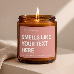 Peut inclure: Un pot de bougie en verre ambré allumé avec une étiquette rose qui dit "SMELLS LIKE YOUR TEXT HERE". La bougie est étiquetée "8oz - Soy Candle - Hand Poured in Canada". La bougie est sur une surface neutre.