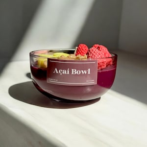 May include: A clear glass bowl-shaped candle with a deep purple wax base, resembling an açai bowl. The candle is topped with wax fruit, including red strawberries and yellow mango slices. The label reads "Açai Bowl" and "Fruity Scent".