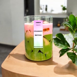 May include: A layered drink in a clear glass, featuring green matcha at the bottom, a pink strawberry layer in the middle, and a garnish on top. A label reads "AMBER TORONTO STRAWBERRY MATCHA". The drink sits on a light wooden surface.