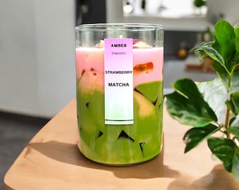 May include: A layered drink in a clear glass, featuring green matcha at the bottom, a pink strawberry layer in the middle, and a garnish on top. A label reads "AMBER TORONTO STRAWBERRY MATCHA". The drink sits on a light wooden surface.