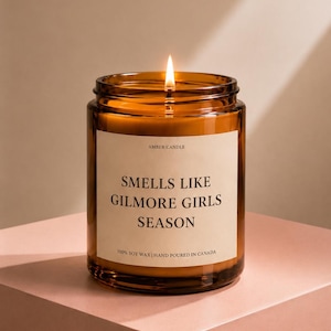 May include: A lit amber glass jar candle with a cream-coloured label that reads AMBER CANDLE, SMELLS LIKE GILMORE GIRLS SEASON, and 100% SOY WAX | HAND POURED IN CANADA. The candle features a single burning wick centred in the wax.
