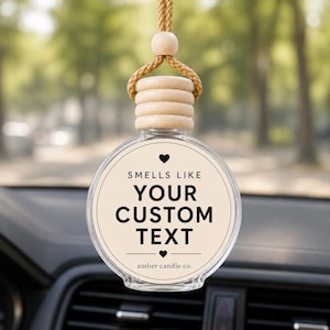 Peut inclure: Un flacon diffuseur de parfum pour voiture en verre transparent rond avec une étiquette beige clair. Elle porte un petit cœur, le texte "SMELLS LIKE YOUR CUSTOM TEXT" et "amber candle co." en bas. Il possède un bouchon en bois rainuré et une cordelette.