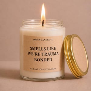 Peut inclure: Une bougie allumée dans un pot en verre transparent avec un couvercle doré. L'étiquette indique AMBER CANDLE CO. SMELLS LIKE WE'RE TRAUMA BONDED 8oz HAND POURED IN CANADA. La cire est blanche et la flamme est visible au sommet de la mèche.