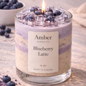 May include: A lit candle in a glass jar with a layered design. The candle is labeled "Blueberry Latte" and topped with blueberries and ice. The jar is 16 oz and made in Canada.