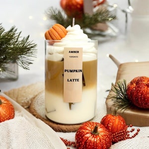 May include: A glass candle with a layered design resembling a pumpkin latte. The candle has a white base, a tan middle, and a whipped cream-like top, topped with a small orange pumpkin. The label reads "AMBER TORONTO PUMPKIN LATTE."