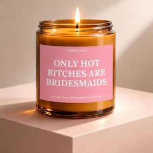 Peut inclure: Une bougie allumée dans un pot en verre ambré avec une étiquette rectangulaire rose portant le texte blanc AMBER CANDLE, ONLY HOT BITCHES ARE BRIDESMAIDS, et 100% SOY WAX | HAND POURED IN CANADA. La cire est de couleur brun doré.