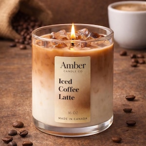 May include: A clear glass candle with a layered design, resembling an iced coffee latte. The candle has a lit wick, ice cubes, and a label that reads "Amber Candle Co. Iced Coffee Latte 16 OZ Made in Canada." Coffee beans are scattered around the candle.
