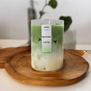 Può includere: Una candela di vetro con un design matcha latte. La candela ha uno strato superiore verde, uno strato intermedio bianco e un'etichetta che dice "AMBER TORONTO MATCHA LATTE". Si trova su un vassoio di legno.