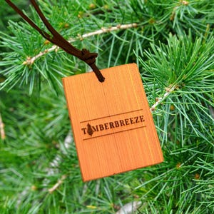 May include: A rectangular wooden tag with the word "TIMBERBREEZE" engraved on it, hanging from a brown cord. The tag is a warm orange color and is set against a backdrop of green pine needles.
