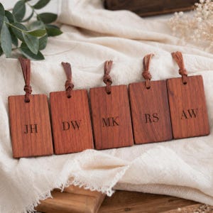 May include: Five rectangular wooden tags with leather loops. Each tag is engraved with initials: JH, DW, MK, RS, and AW. The tags are a warm brown color and appear to be made of hardwood.