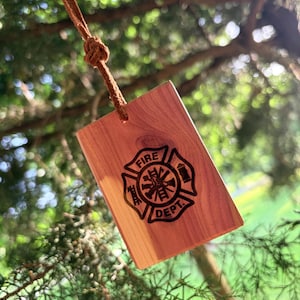 May include: A rectangular cedar wood ornament featuring a laser-engraved black Fire Dept emblem with a Maltese cross design. The piece is suspended by a thin brown leather cord tied in a knot at the top, showcasing the natural wood grain texture.