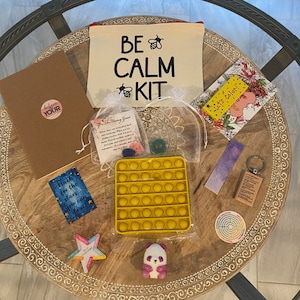 Puede incluir: Una bolsa de lona beige con el texto "BE CALM KIT" en letras negras y un gráfico de abeja. La bolsa está rodeada de varios artículos, incluyendo una libreta marrón con el texto "Believe Your" en letras blancas, un juguete de burbujas amarillo, un pequeño peluche de panda, un adorno de madera en forma de estrella, una pequeña caja de madera con una llave adjunta, un marcapáginas morado y blanco, una pequeña tarjeta con el texto "Reach for the stars you can do it!" en letras azules, una pequeña tarjeta con el texto "Let's Color!" en letras amarillas, y una pequeña tarjeta con un diseño floral.