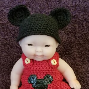 Puede incluir: Una muñeca de crochet con un gorro negro de punto con orejas, un atuendo de crochet Mickey Mouse rojo con botones amarillos y zapatos de crochet amarillos. La muñeca está sentada sobre una superficie de felpa morada. Sweet Silver Creations 2019 está escrito debajo de la muñeca.