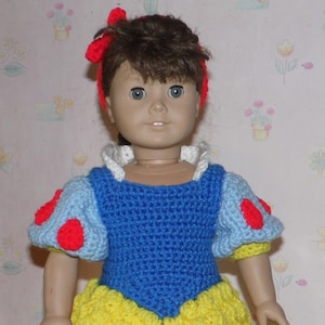 May include: A crocheted Snow White costume for a doll, featuring a blue bodice with red accents, a white collar, and a yellow skirt. The doll is wearing a red headband with a bow.