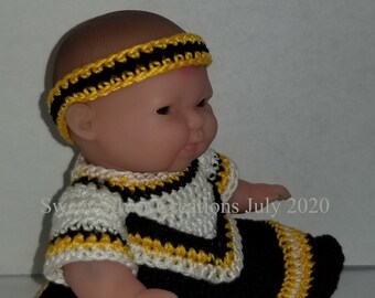 Puede incluir: Una muñeca pequeña con un atuendo de ganchillo negro y blanco con ribetes amarillos. El atuendo incluye un vestido, polainas y una diadema.