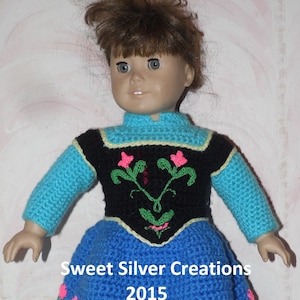 May include: A crocheted doll dress in blue, black, and green with pink flowers. The dress has a black bodice with a floral design and a blue skirt with a green and pink floral trim. The dress is made for a doll with brown hair and blue eyes. The text "Sweet Silver Creations 2015" is visible on the dress.