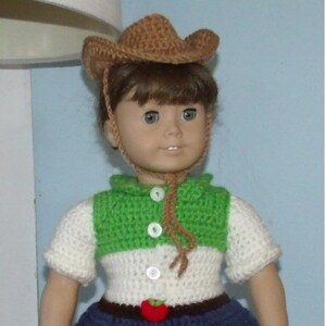 Puede incluir: Una muñeca con un atuendo de ganchillo con una falda verde, blanca y azul, un sombrero de vaquero marrón y botas marrones con detalles de manzanas rojas. La muñeca sostiene una manzana roja en su mano. El texto "Sweet Silver Creations 2016" es visible en la falda.