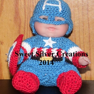 Könnte beinhalten: Eine gehäkelte Captain America Puppe, die ein blaues, weißes und rotes Kostüm mit einem weißen Stern auf der Brust trägt. Die Puppe sitzt auf einer Holzoberfläche. Sweet Silver Creations 2014.