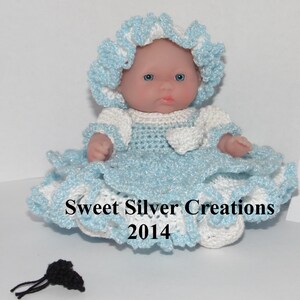 Könnte beinhalten: Eine kleine, gehäkelte Puppe, die ein blaues und weißes Kleid und einen Hut trägt. Die Puppe hat blaue Augen und ein rosafarbenes Gesicht. Die Puppe sitzt auf einer weißen Oberfläche. Sweet Silver Creations 2014.