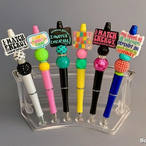 May include: A collection of decorative pens with various colors and designs. Each pen features a unique topper with phrases like "I match energy" and "My level of sarcasm depends on your level of stupidity."