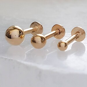 May include: Three gold-colored barbell earrings with round balls on each end. The earrings are different sizes.