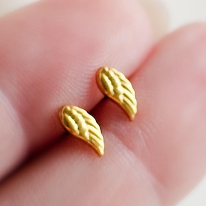 May include: A pair of small, gold-coloured wing-shaped stud earrings. The earrings have a textured surface, and are displayed against a blurred background.